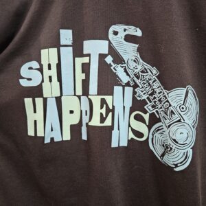 Blue Shirt with text that reads Shift Happens in blue and teal ink. There is also a woodcut block print of an old campagnolo derailleur beside the text