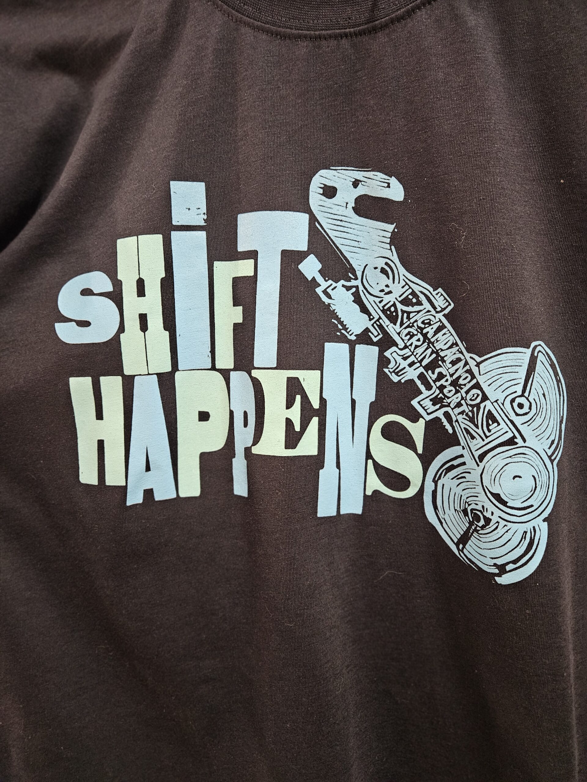 Blue Shirt with text that reads Shift Happens in blue and teal ink. There is also a woodcut block print of an old campagnolo derailleur beside the text