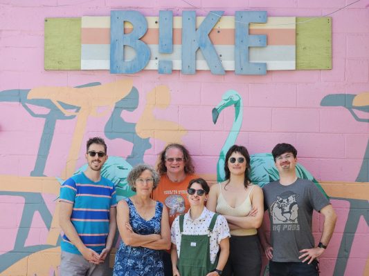 This is a photo of the staff team. Moving right to left, there is Mark, Jean, Lily, Ness, Tahirih, Erik. They are posed against the flamingo mural in the back alley.